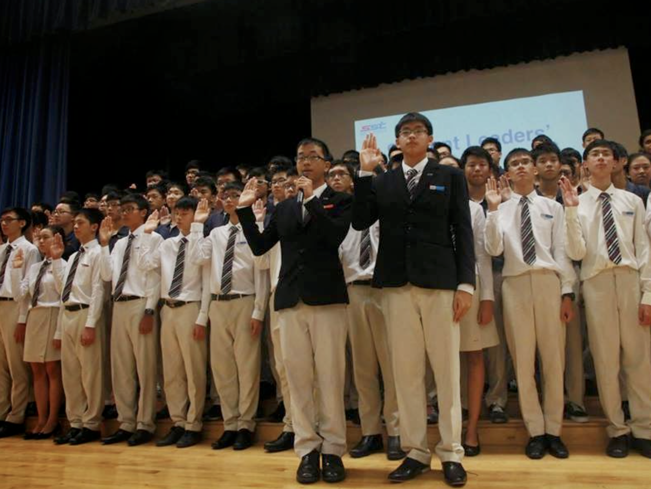 A large group of uniformed students raise their right hands on a stage during what appears to be a pledge ceremony.